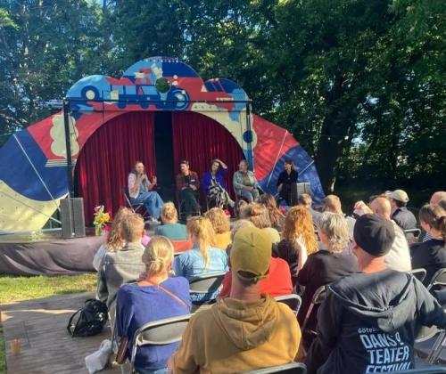 publiek op stoelen kijkt in buitenlucht naar drie sprekers op een klein podium en decor